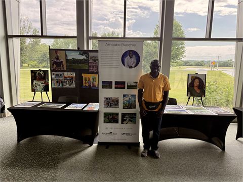Amoako Buachie standing beside his display table of prints and biography panels at Archipelago 2026 in Fond du Lac, Wisconsin.