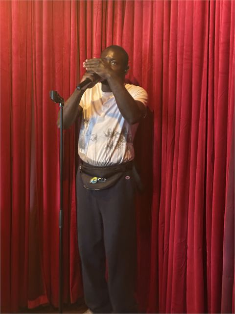 Amoako Buachie standing on a small stage in front of a red velvet curtain at a karaoke lounge.